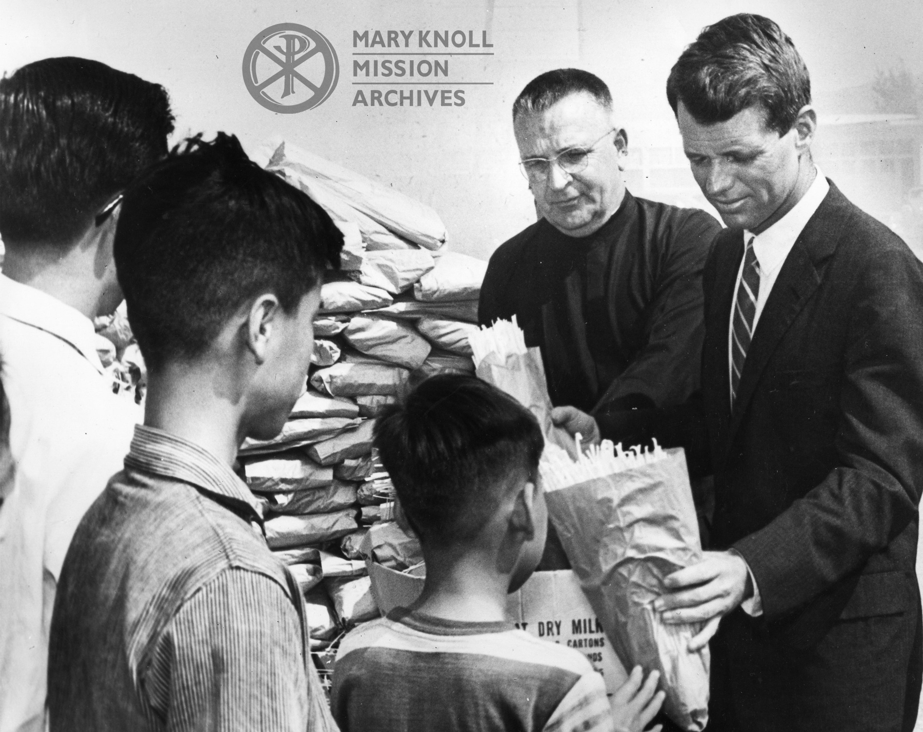 Robert Kennedy distributes food with Fr. Howard Trube, MM at the Bishop Ford Memorial Center in Hong Kong, February 12, 1962