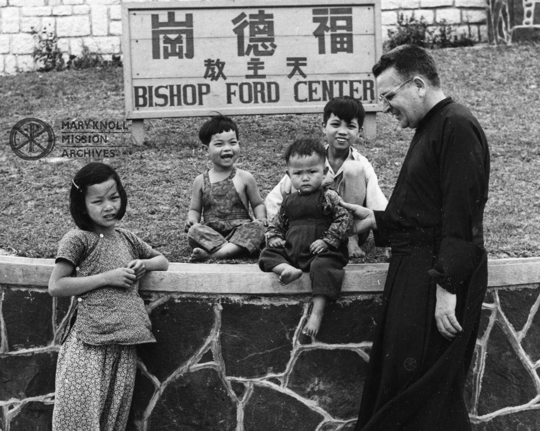 Fr. Trube with Chinese Children in front of the Bishop Ford Center sign, c. 1953